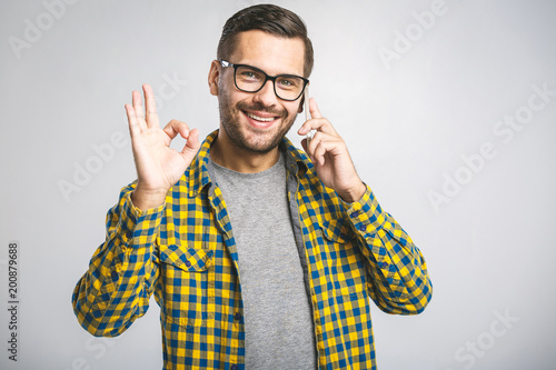 Smiling young casual man talking on the phone and making ok sign on the grey background.