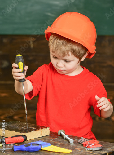 Kid boy holds screwdriver tool. Toddler on busy face plays with screwdriver at workshop. Handcrafting and workshop concept. Child in helmet cute playing as builder or repairer, or handcrafting.