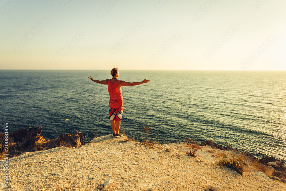 A man is practicing yoga on the shore of a rocky ocean. The young man opened his hands and looked out into the distance to the sea.