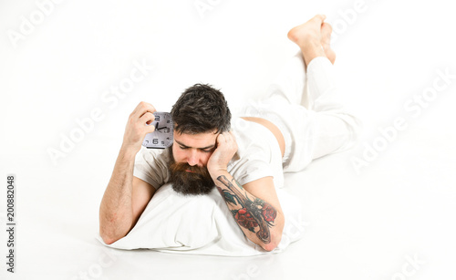 Guy with sleepy face lies on pillow, holds alarm clock.