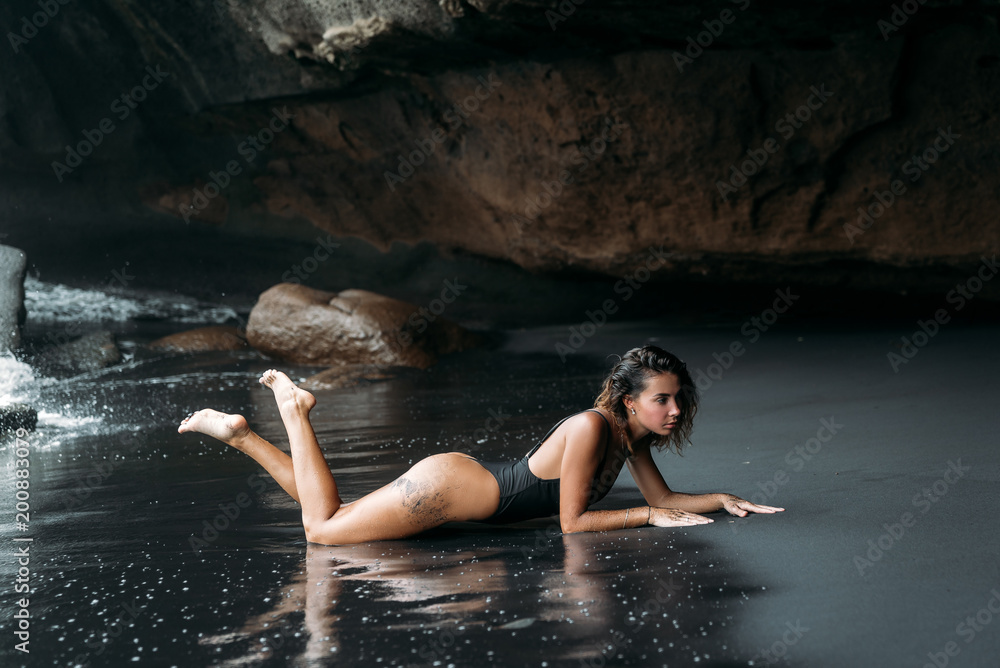 Fototapeta premium Beautiful girl with sexy body in black swimsuit lying on the beach with black volcanic sand. Young model with curly hairs, tanned skin posing and relaxing in the journey.