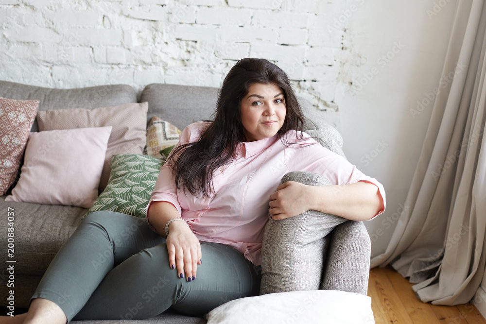 Joy, rest, relaxation and happiness concept. Picture of body positive attractive 25 year old woman with big breast and hips spending leisure time indoors, sitting comfortably on gray couch and smiling Joy, rest, relaxation and happiness concept. Picture of body positive attractive 25 year old woman with big breast and hips spending leisure time indoors, sitting comfortably on gray couch and smiling