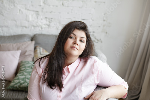Close up of attractive chubby girl in her twenties resting at home after she finished doing housework, sitting on couch in living room, thinking about something. People, rest, leisure and relaxation