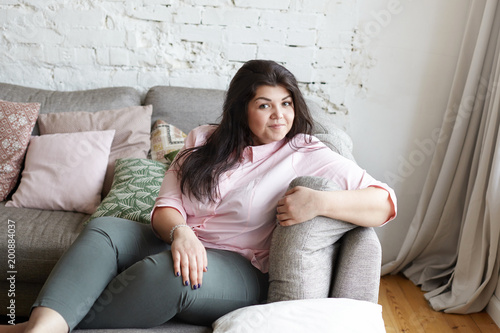 Joy, rest, relaxation and happiness concept. Picture of body positive attractive 25 year old woman with big breast and hips spending leisure time indoors, sitting comfortably on gray couch and smiling