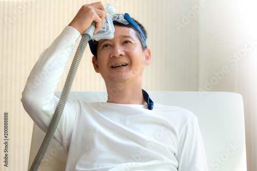 Sleep apnea therapy. Man laying in bed holding CPAP mask with a smiling face after waking up from daytime sleepiness. Elderly patient of obstructive sleep apnea feels fresh with CPAP machine therapy.