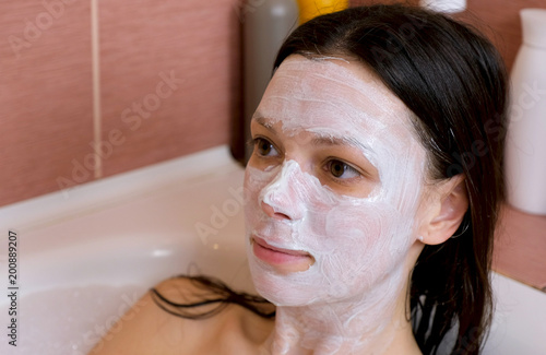Beautiful brunette woman with blue clay mask on her face in the bathroom. Spa at home. Close-up face.