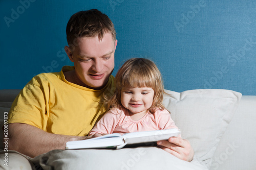 Father reading a story to his child girl sitting on couch at home. Happy family time together at home. 