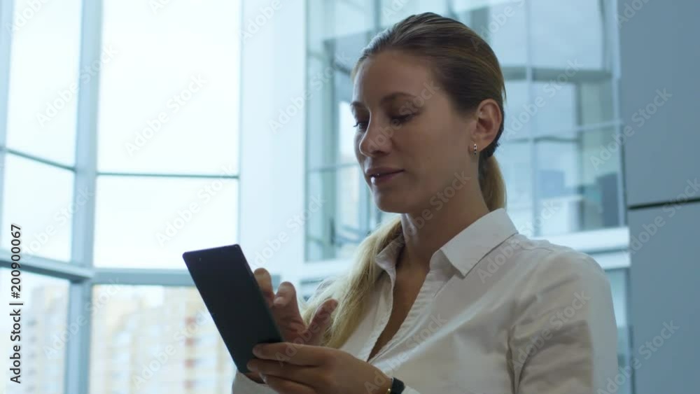 Panning shot of mid-aged attractive businesswoman with blonde hair standing in office center and using smart phone Panning shot of mid-aged attractive businesswoman with blonde hair standing in office center and using smart phone