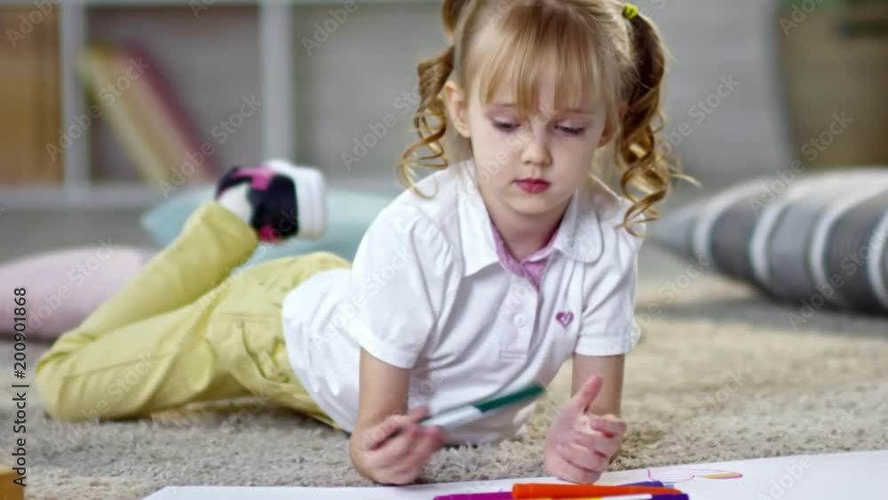 Adorable little girl with wavy golden hair and two ponytails lying on the floor and using colored felt-tip pens to draw a picture Adorable little girl with wavy golden hair and two ponytails lying on the floor and using colored felt-tip pens to draw a picture
