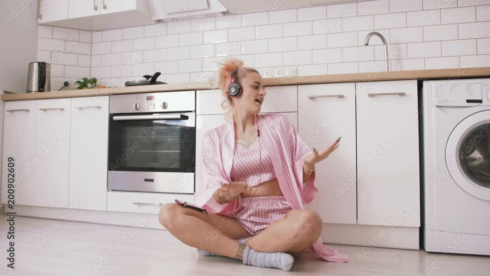 Happy young woman with pink hair sitting on the floor on kitchen wearing pink pajamas and listening to music with headphones Happy young woman with pink hair sitting on the floor on kitchen wearing pink pajamas and listening to music with headphones