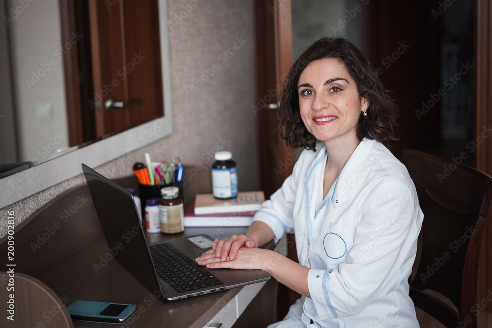 A young smiling brunette doctor woman in white medical gownsits at her desk, which has a laptop, books, accessories and jars of medicines or vitamins. A young smiling brunette doctor woman in white medical gownsits at her desk, which has a laptop, books, accessories and jars of medicines or vitamins.
