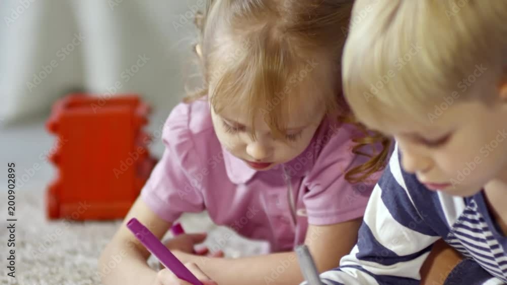 Two little children, girl and boy, lying on the floor and drawing picture together using colored felt-tip pens Two little children, girl and boy, lying on the floor and drawing picture together using colored felt-tip pens