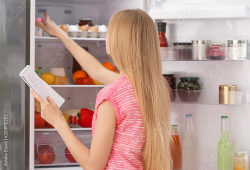 Young woman with notebook near open refrigerator in kitchen