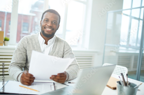 Waist-up portrait of smiling African American manager looking at camera while sitting at office desk and finishing promising project  interior of modern office on background