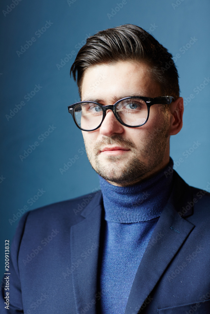 Attractive confident Caucasian man wearing turtleneck, jacket and glasses looking at camera on blue background Attractive confident Caucasian man wearing turtleneck, jacket and glasses looking at camera on blue background