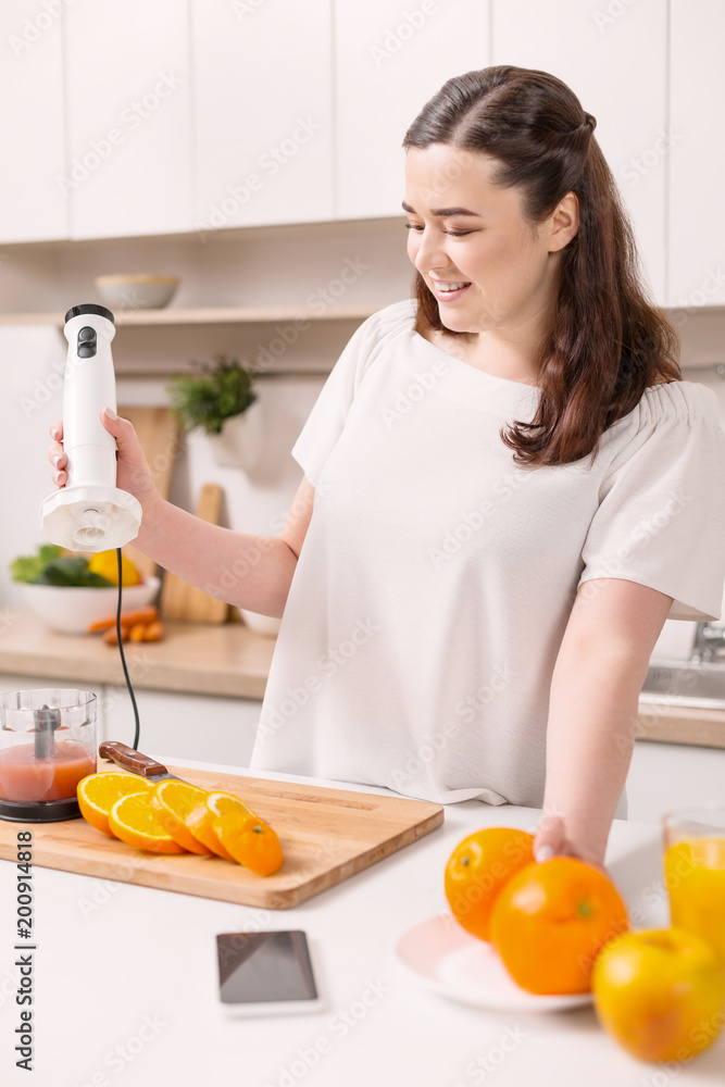 Full of vitamins. Thoughtful positive woman looking down while carrying blender