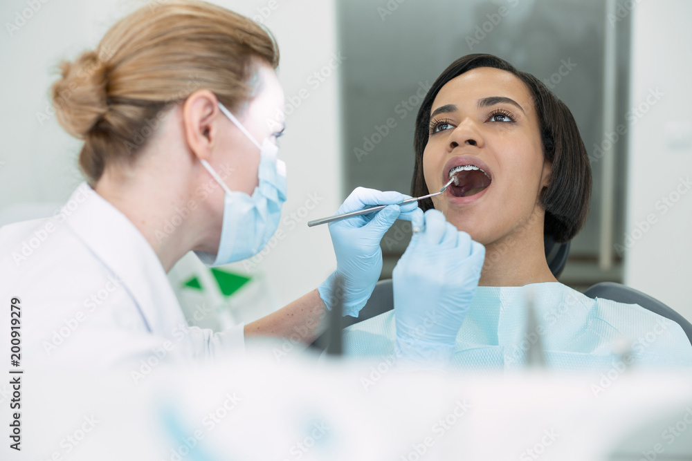 Visiting the doctor. Unsmiling dark-haired patient sitting with her mouth opened while the dentist examining her teeth Visiting the doctor. Unsmiling dark-haired patient sitting with her mouth opened while the dentist examining her teeth