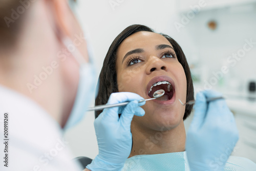 Visiting the dentist. Concentrated dark-haired patient sitting with her mouth opened while the dentist examining her teeth