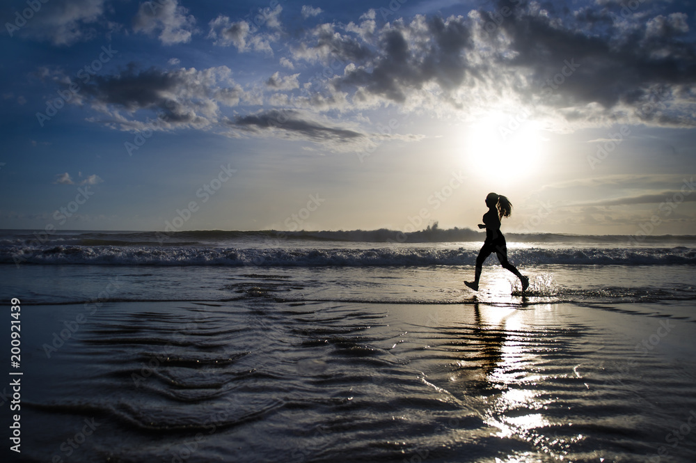 silhouette of young Asian sport runner woman in running workout training at sunset beach with orange sunlight reflection on the sea water