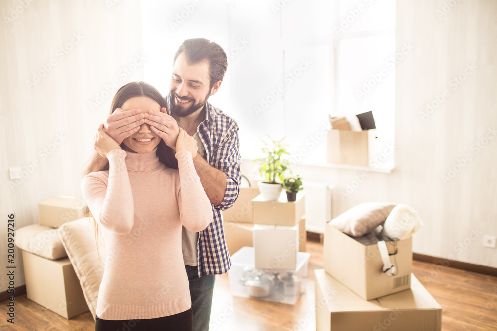 Happy couple are standing inside their new house. Young man has closed eyes to his wife. He prepared surprise for her. She is holding her hands on his. Happy couple are standing inside their new house. Young man has closed eyes to his wife. He prepared surprise for her. She is holding her hands on his.