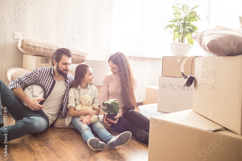 Two young parents are sitting on the floor and looking to their child. They are surrounded with oxes full of stuff for kitchen and living room. They are spending time with each other and talking.