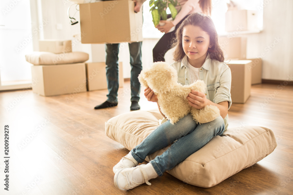 Small girl is sitting on the pillow on the floor and looking to her teddy bear toy. She has some rest while her parents are holding boxes with things and moving them to another parts of apartment. Small girl is sitting on the pillow on the floor and looking to her teddy bear toy. She has some rest while her parents are holding boxes with things and moving them to another parts of apartment.