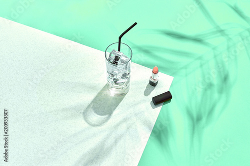 High angle view of lipstick and drinking glass with ice on table. Blue and white background with shadow from a palm leaf photo