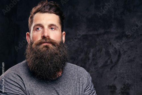 Portrait of a hipster with full beard and stylish haircut, dressed in a gray t-shirt, stands with a thinking look in a studio on a dark background.