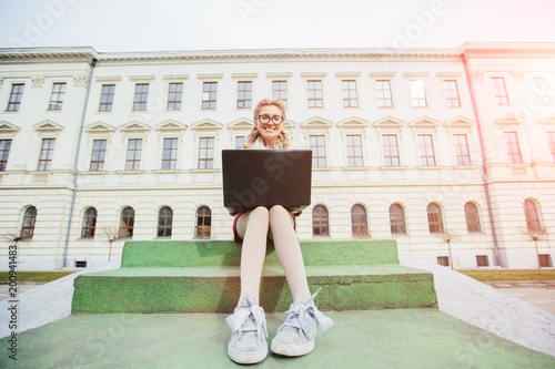 Young beautiful excited student woman smiling and looking at camera with laptop sitting on stairs near the university buiding background. Wide angle photo