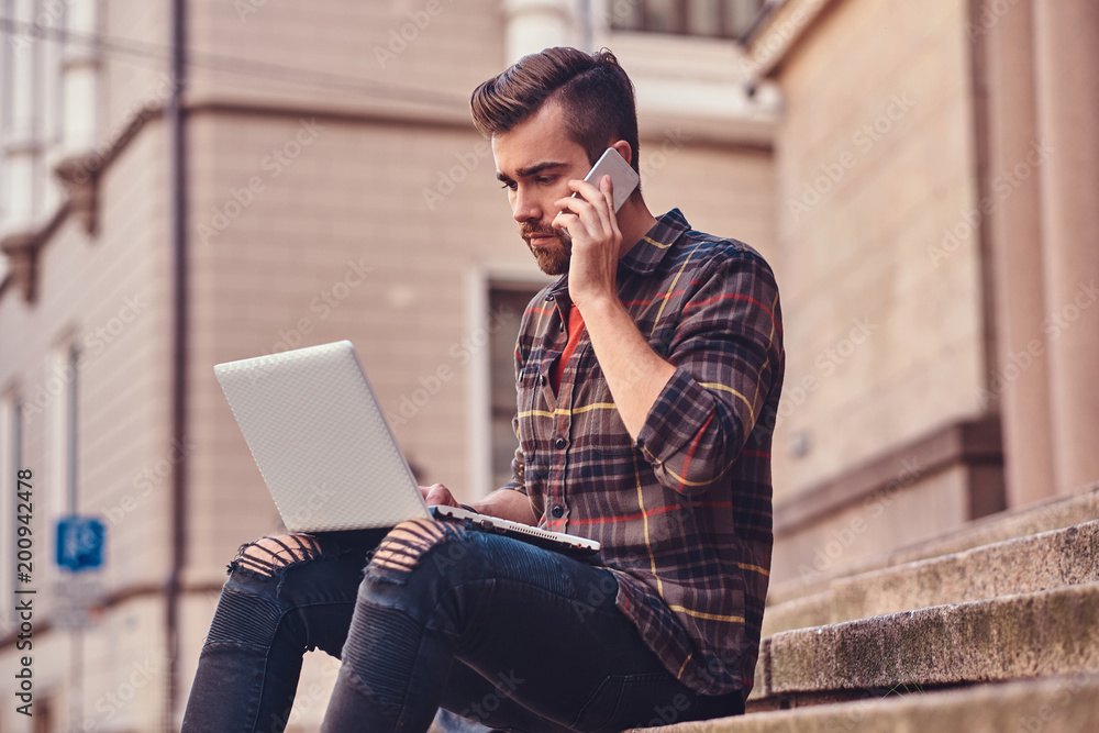 Beautiful male with stylish haircut and beard, wearing a fleece shirt and jeans, sitting on steps against an old building. Beautiful male with stylish haircut and beard, wearing a fleece shirt and jeans, sitting on steps against an old building.