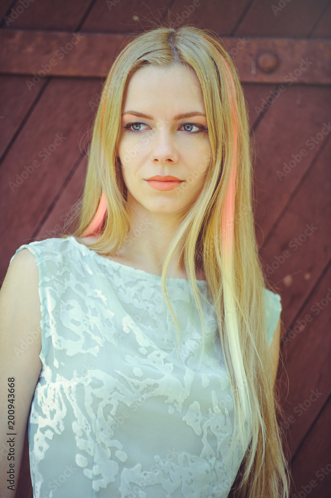 Attractive modest young blond woman on red wooden background her hair painted in pink striped striped, in blue dress shy and inquisitive Attractive modest young blond woman on red wooden background her hair painted in pink striped striped, in blue dress shy and inquisitive