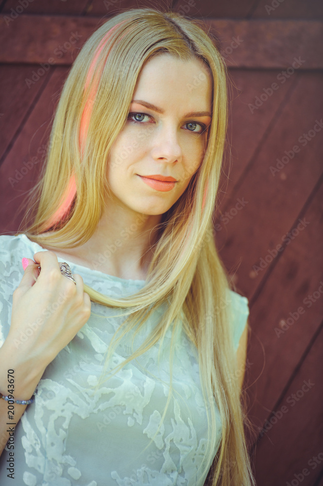 Fototapeta premium Attractive modest young blond woman on red wooden background her hair painted in pink striped striped, in blue dress shy and inquisitive