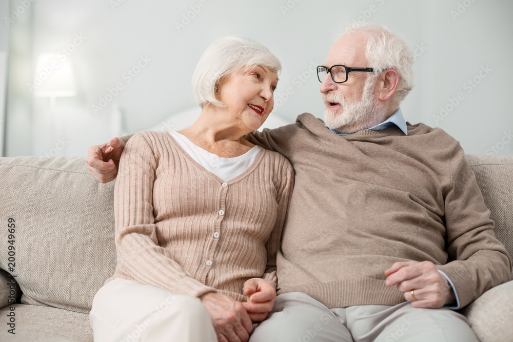 Pleasurable rest. Positive nice aged man sitting on the sofa while talking to his wife