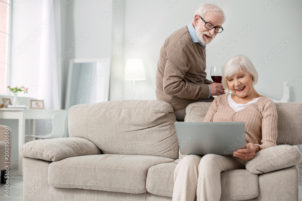 Modern device. Delighted aged woman sitting on the sofa while using her laptop Modern device. Delighted aged woman sitting on the sofa while using her laptop
