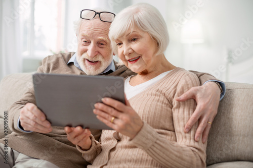 Modern tablet. Nice pleasant senior couple sitting together on the sofa while using modern tablet