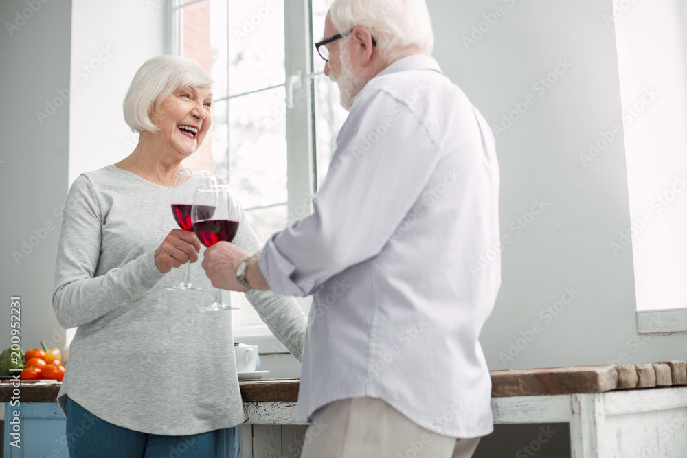 Fototapeta premium Pleasant celebration. Delighted happy senior couple smiling to each other while clinking glasses with wine