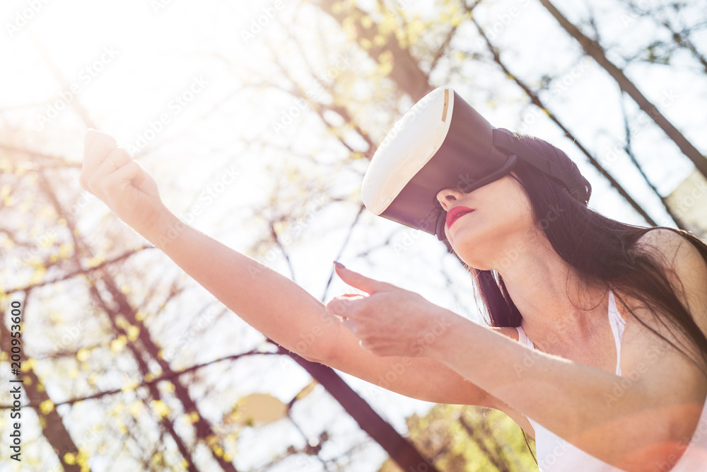 Naklejka premium Virtual reality, fun in the park. A woman relaxes in the park using googles VR. The girl imagines that she is climbing the rope. Spending free time outside, lifestyle concept.