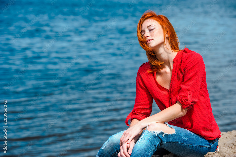 Joyful redhead woman sitting comfortably, feeling serene and free and enjoying a sunny day at the beach.