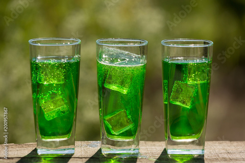 three glasses with an exotic green carbonated drink with ice on the board, against a background of greenery