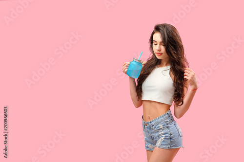 Young slim brunette in denim shorts and top having refreshing drink posing flirty on pink backdrop.