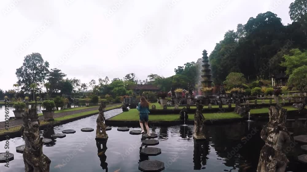 Camera goes up: young woman runs along tiles in a pond in a beautiful Balinese temple Tirta Gangga in Bali, slow motion Camera goes up: young woman runs along tiles in a pond in a beautiful Balinese temple Tirta Gangga in Bali, slow motion