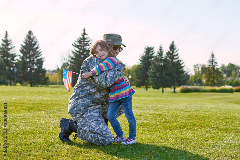 Obraz premium Soldier reunited with his daughter. Child with usa flag is hugging her military daddy outdoor in the summer park.