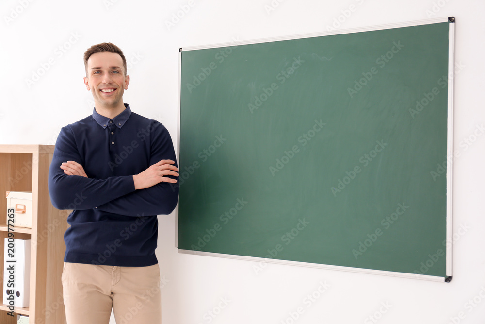 Young male teacher standing near blackboard in classroom Young male teacher standing near blackboard in classroom