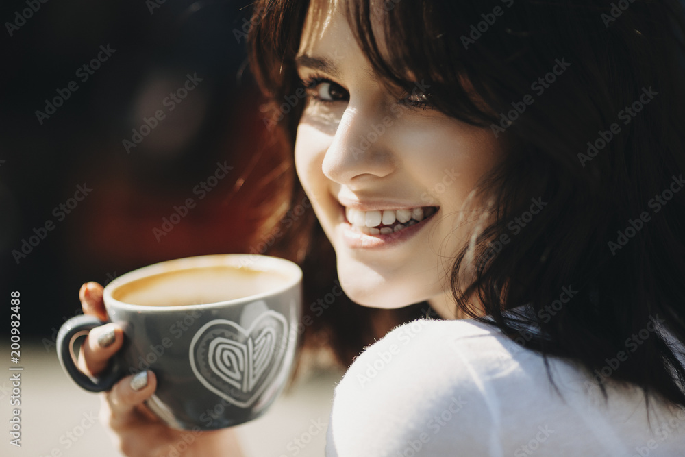 Close up portrait of a beautiful young woman that is holding a cup of cappucino in hand and is smilling at camera.