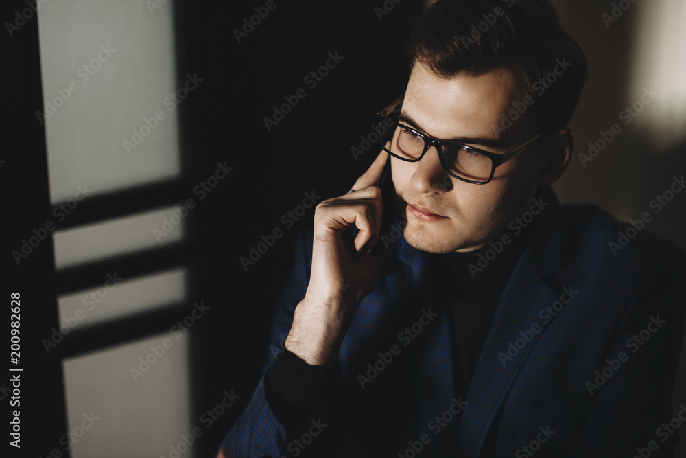 Portrait of serious businessman with glasses sitting on a chair and looking away while speaking on the phone in a dark room Portrait of serious businessman with glasses sitting on a chair and looking away while speaking on the phone in a dark room