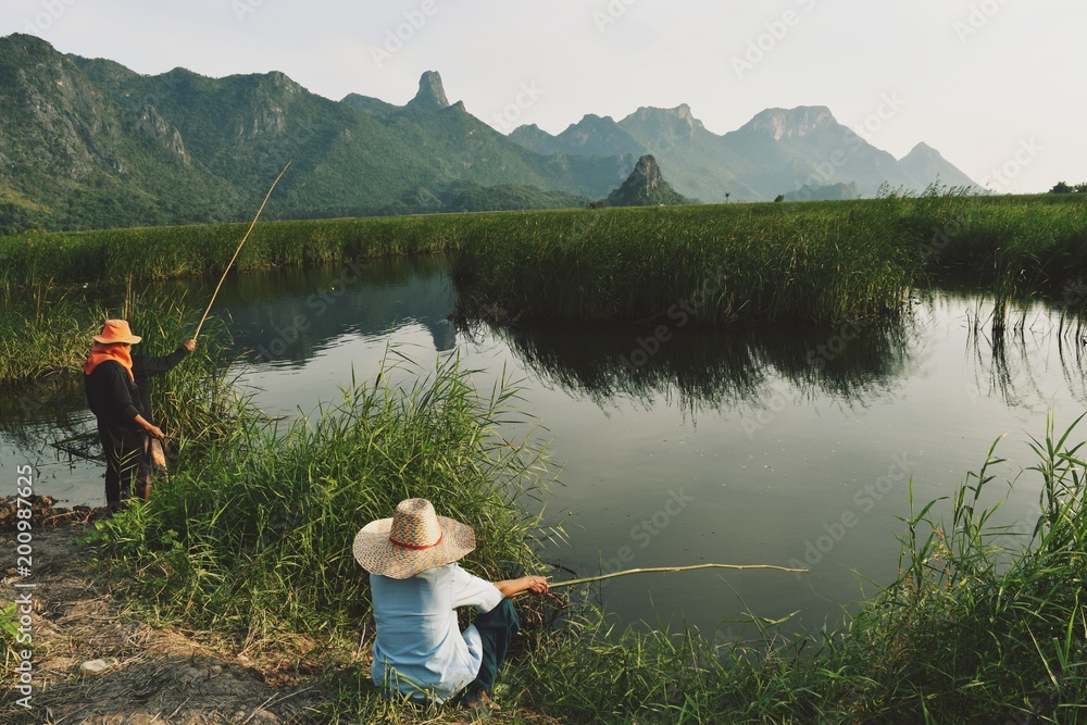 Fisherman at stream in the front of mountain, People wear hat and holding  bamboo fishing rod, Beautiful view of tranquil water reflecting the  limestone mountain, Thailand Stock Photo