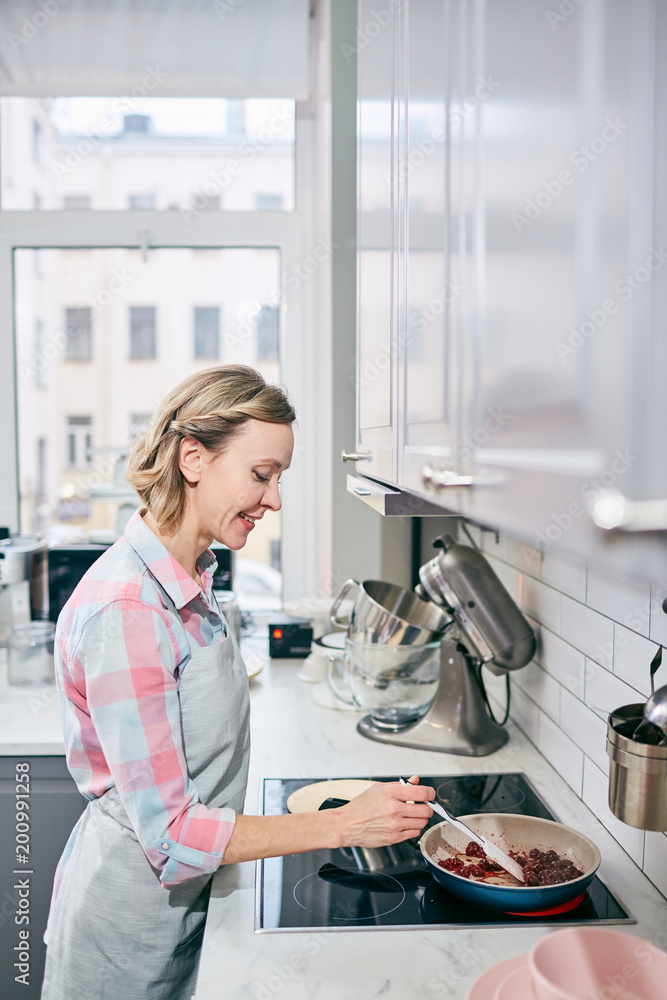 Side view of good-looking Caucasian woman preparing meal in frying pan in modern kitchen and smiling happily