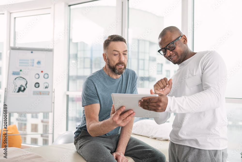 Video lesson. Joyful nice doctor standing near his patient and showing his the video on the tablet Video lesson. Joyful nice doctor standing near his patient and showing his the video on the tablet
