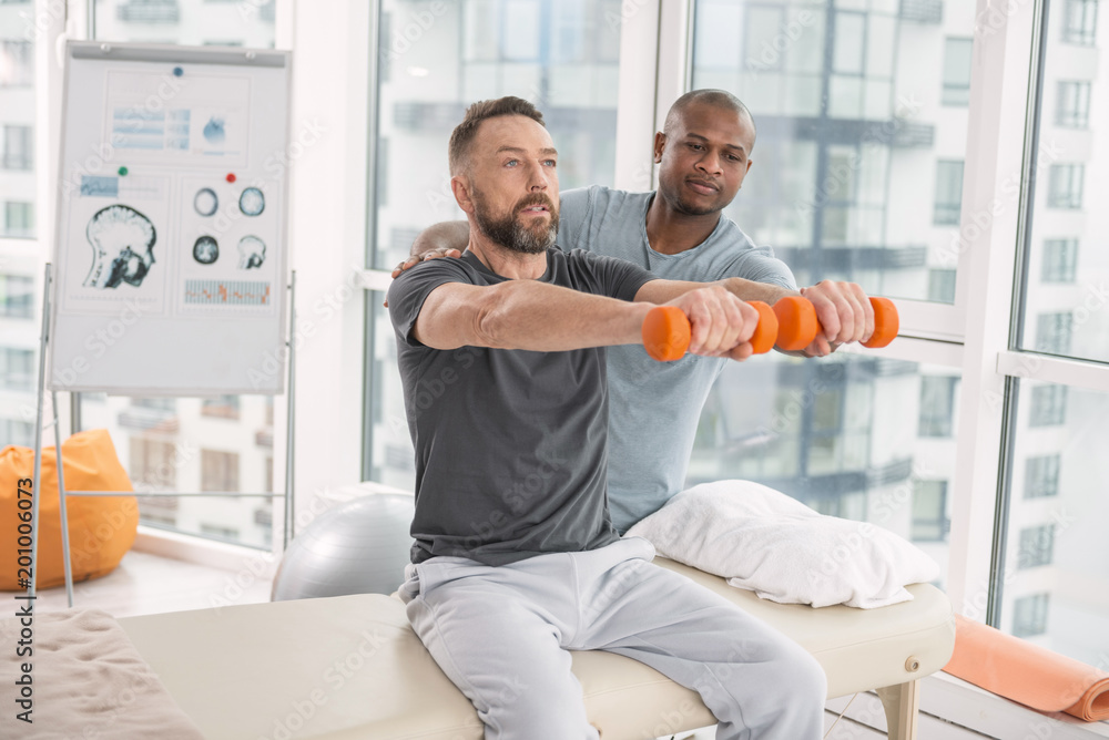 Obraz premium Rehabilitation exercise. Nice pleasant handsome man sitting on the medical couch and holding dumbbells in front of him