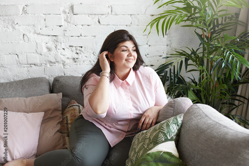 Cute young chubby brunette woman dressed casually having rest on large gray  sofa, smiling shyly, adjusting her dark hair. Portrait of plus size body  positive female enjoying leisure time indoors Stock Photo